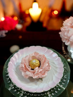 Pink flower with a gold Ferrero Rocher chocolate on a decorative plate with blurred candles and flowers in the background.