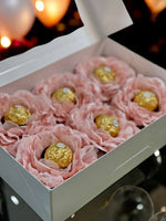 Box of blush flowers holding chocolates wrapped in gold with blurred lights in the background