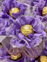 Decorative arrangement with purple flowers and gold Ferrero Rocher chocolates on a white lace background.