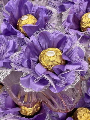 Decorative arrangement with purple flowers and gold Ferrero Rocher chocolates on a white lace background.