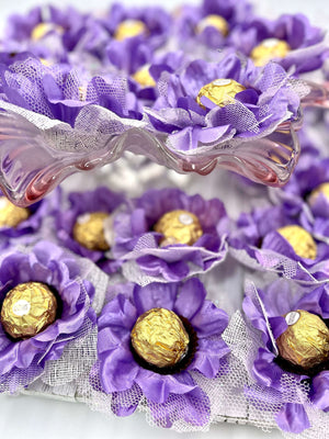Decorative arrangement of purple flowers with gold Ferrero Rocher chocolates inside, on a white background.