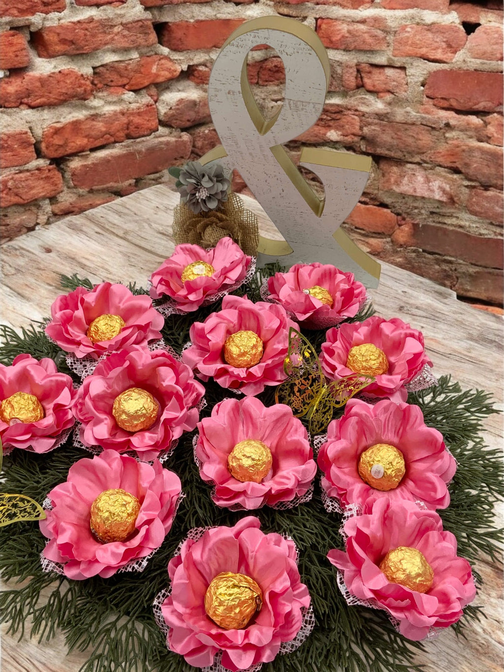 A decorative arrangement of pink silk flowers with gold-colored chocolate holders, presented on a greenery base with a '&' shaped decorative piece above.