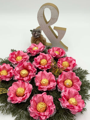 A decorative arrangement of pink silk flowers with gold-colored chocolate holders, presented on a greenery base with a '&' shaped decorative piece above.