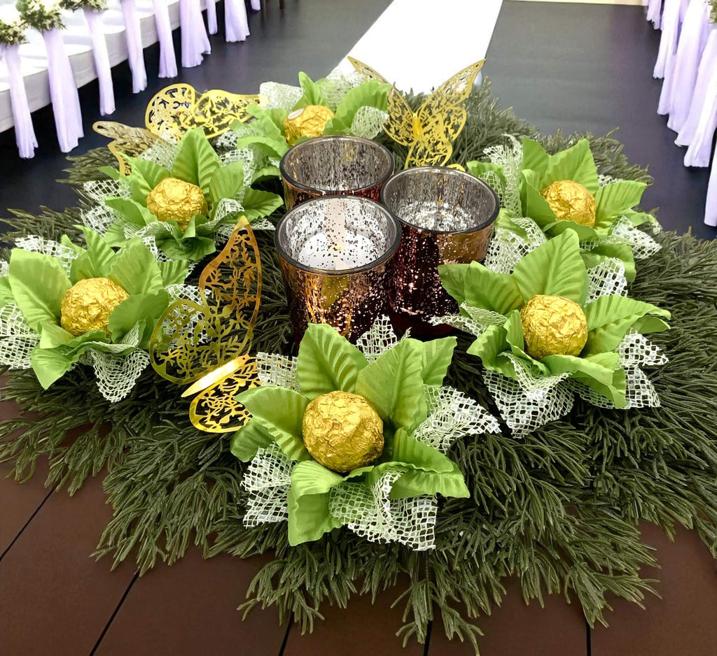 Decorative table centerpiece with green flowers holding chocolate wrapped gold balls, and metallic containers on a wooden table.