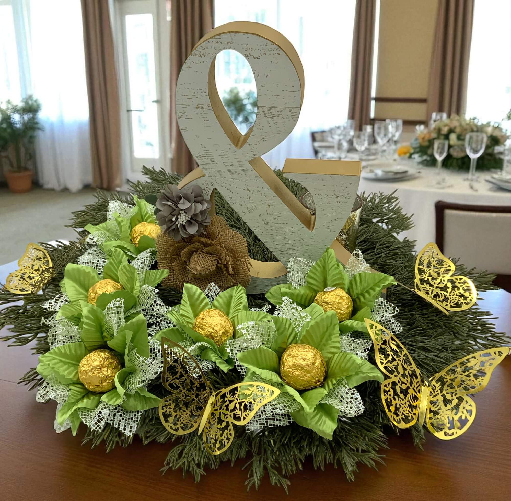 Decorative centerpiece with green fabric flowers chocolate holders, greenery, gold chocolate balls inside each flower, and a large ampersand on a table.