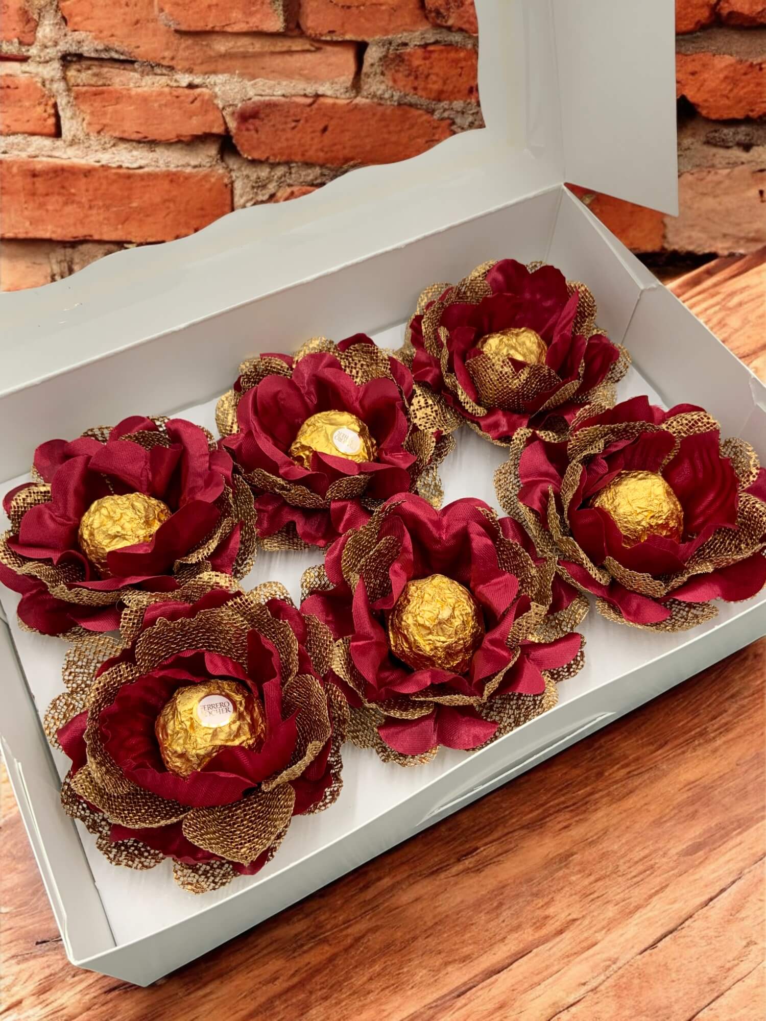 Decorative red and gold flowers with chocolate eggs inside a white box on a wooden surface.