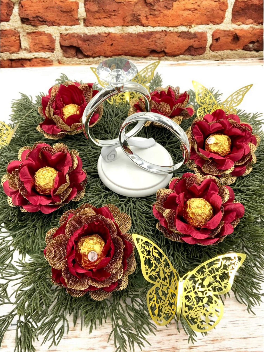 Decorative centerpiece with red and gold flowers holding a gold chocolate and butterfly accents, featuring a ring on a white stand.
