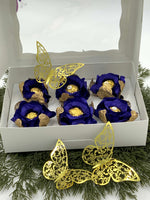 Decorative gift set with blue and gold flowers in a white box, surrounded by greenery.
