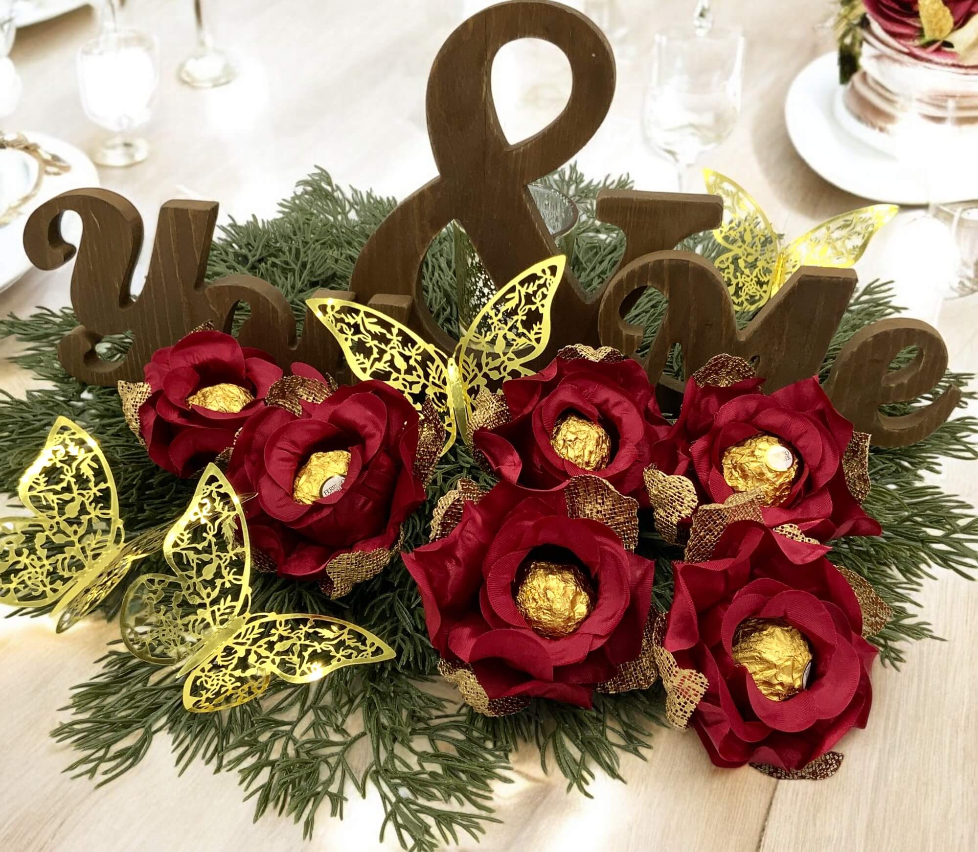 Decorative arrangement with red flowers, gold butterflies, and wooden letters spelling 'You & Me' on a white background.