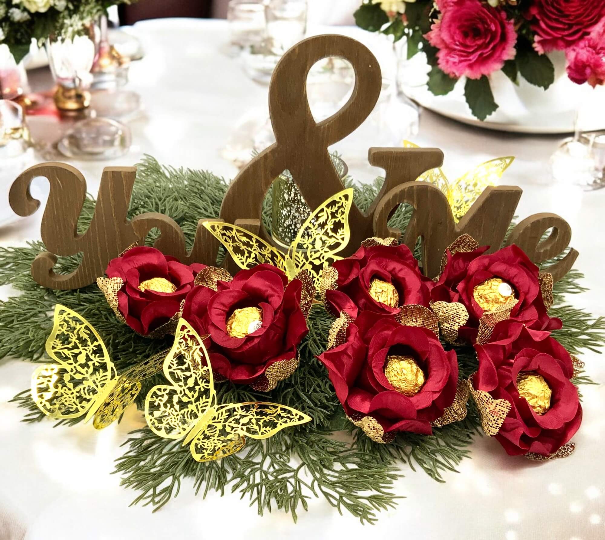 Decorative table centerpiece with red flowers, gold butterflies, and wooden letters on a white tablecloth.