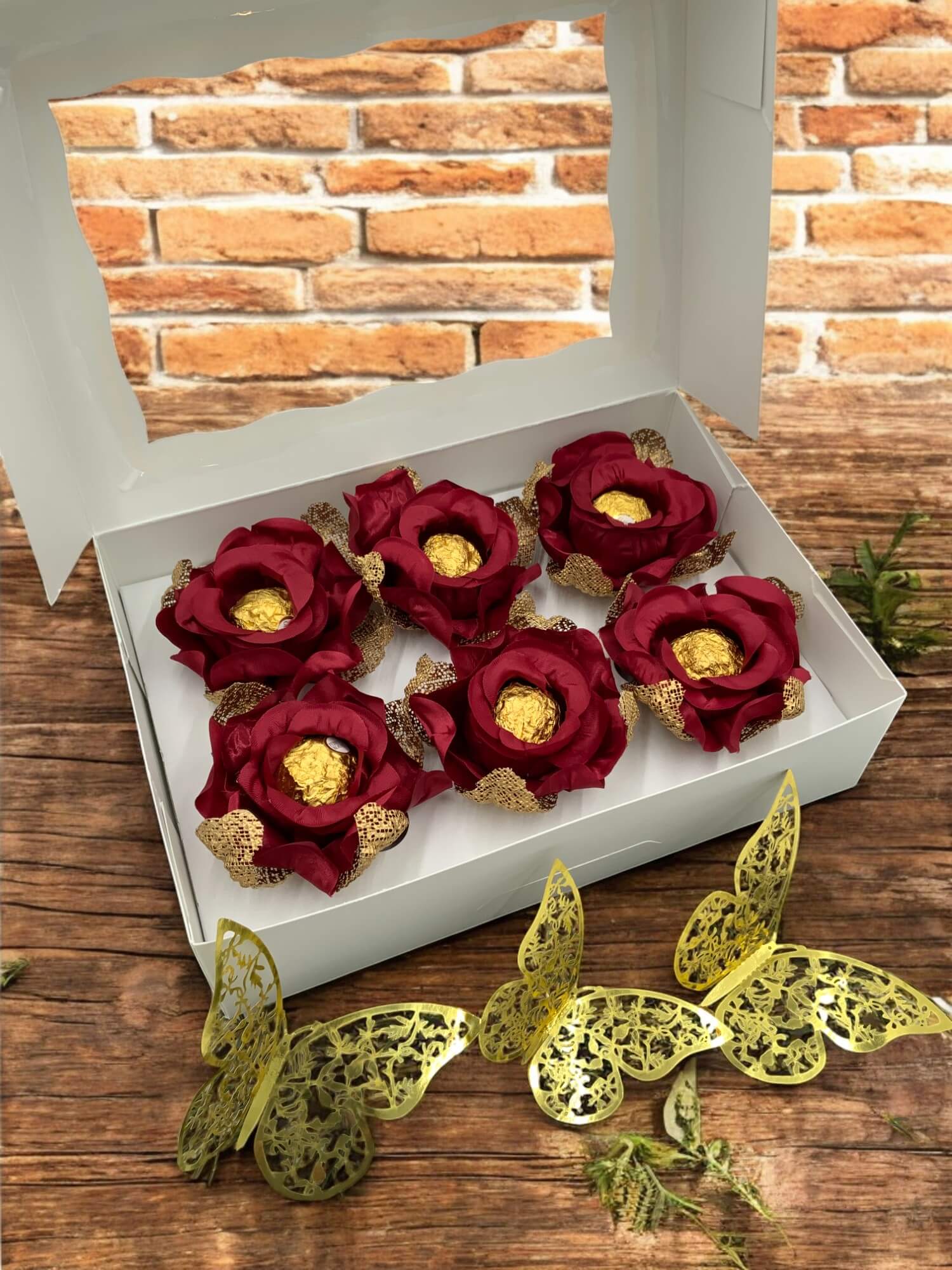 Red floral arrangements with gold chocolate inside them in a white box on a wooden surface with decorative butterflies.