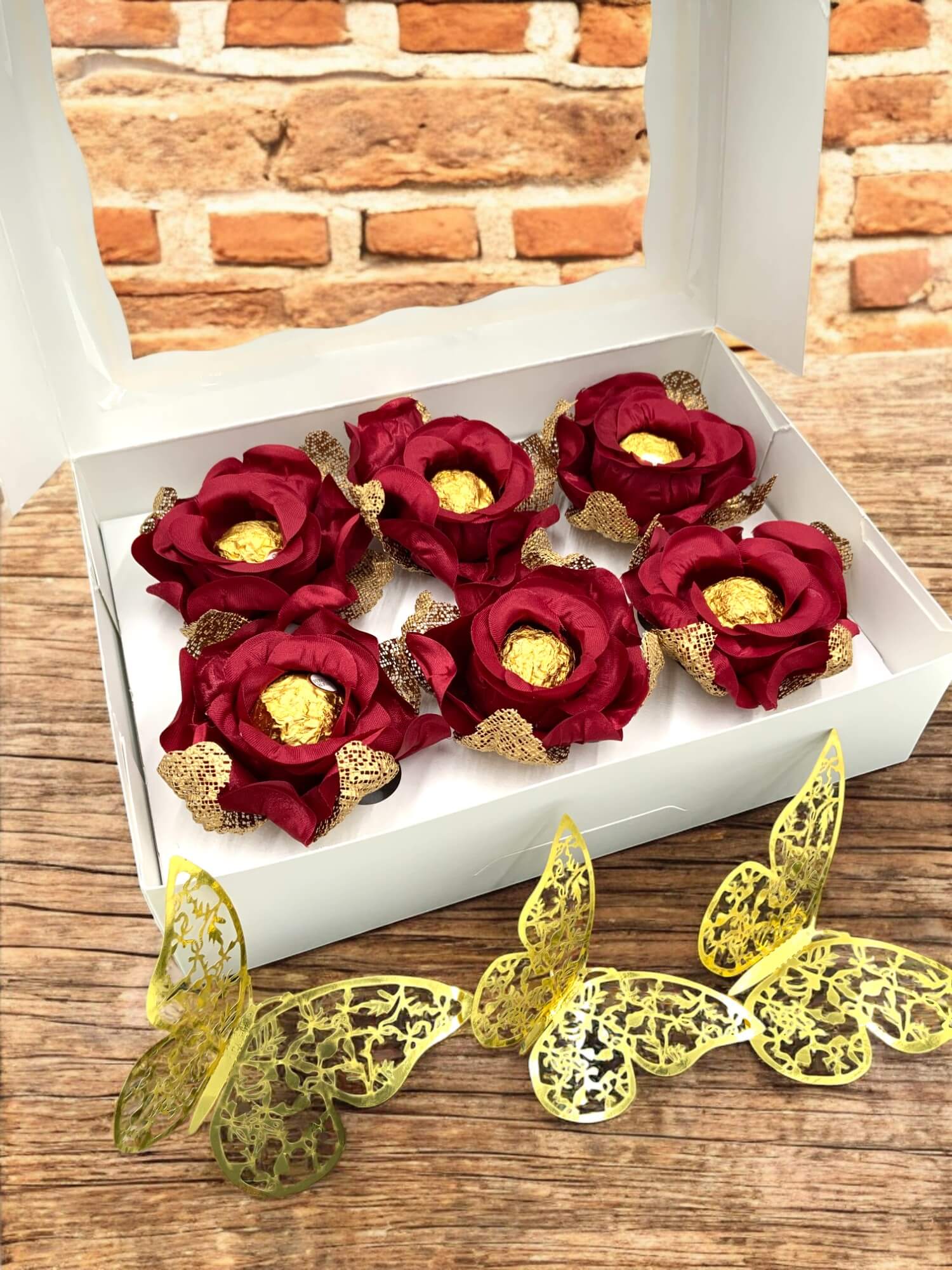 Red flowers with gold chocolate inside them in a white box on a wooden surface with decorative yellow butterflies.