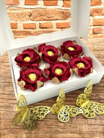 Red flowers with gold chocolate inside them in a white box on a wooden surface with decorative yellow butterflies.