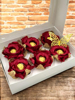 Red flowers with gold centers and decorative butterflies in a white box on a wooden surface.