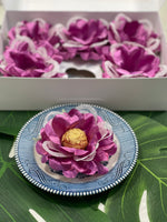 The image show a violet flower with white mesh sittin on a blue saucer. The flower holds a golden chocolate. In the back, there is a box, opened with 5 flowers.
