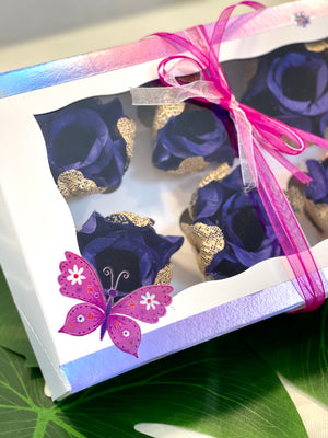The image shows a box of 6 royal blue and gold rosebuds. The box is decorated with a pink butterfly and flowers. there is also a sticker saying "Home is where your mom is". There is a lace made of pink and white bowl made of ribbon.