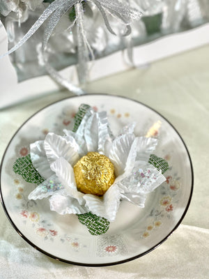 image of a white flower sitting on a saucer. There is a chocolate truffle wrapped in gold foil in the flower