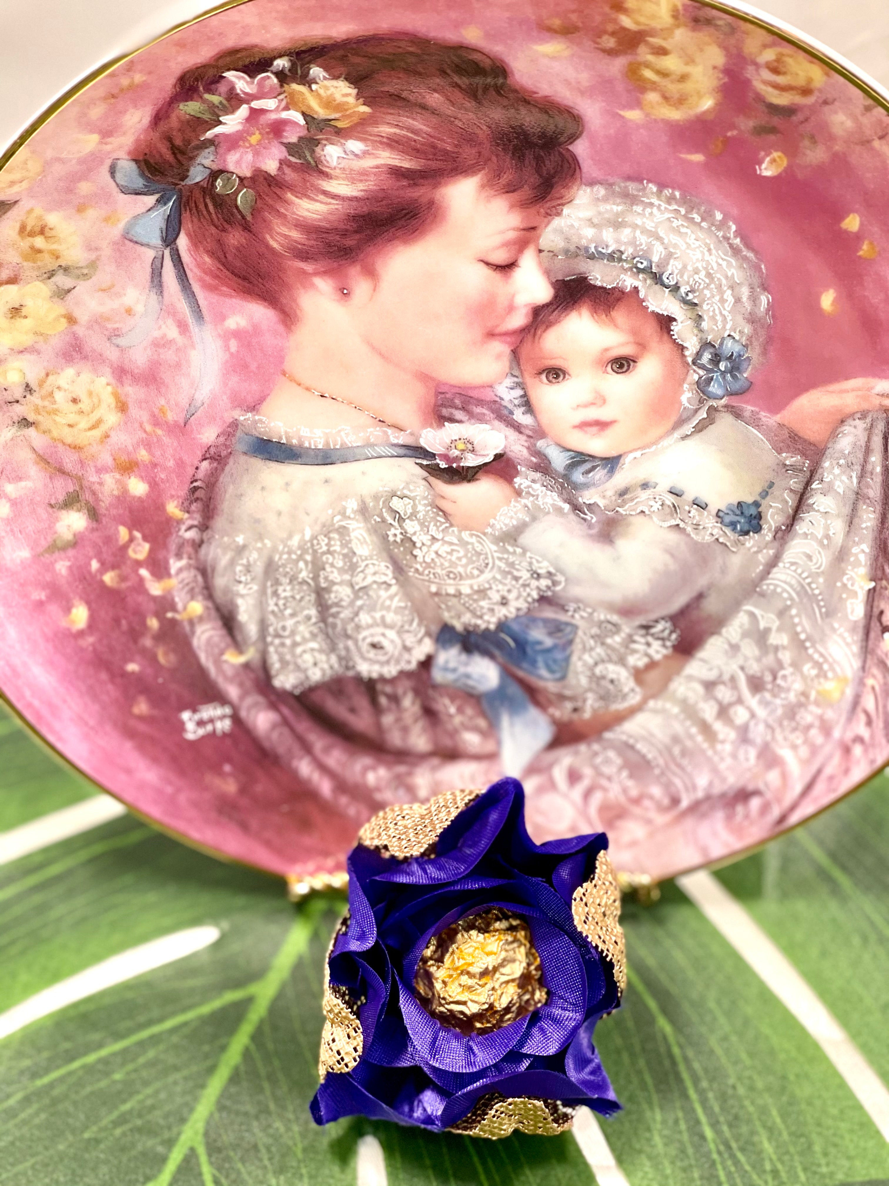 The image shows a decorative plate with a picture of a mother holding a child. In front of the plate sits a rosebud made of royal blue fabric with gold mesh. In the rose there is a chocolate truffle.