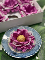 The image show a violet flower with white mesh sittin on a blue saucer. The flower holds a golden chocolate. In the back, there is a box, opened with 5 flowers.