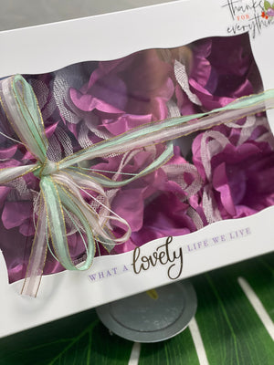 White box decorated with letters saying " What a lovely life we live" and also "thanks for everything". There are 6 violet color flowers in it. It has a lace made of light pink and light green ribbon to adorn the box.