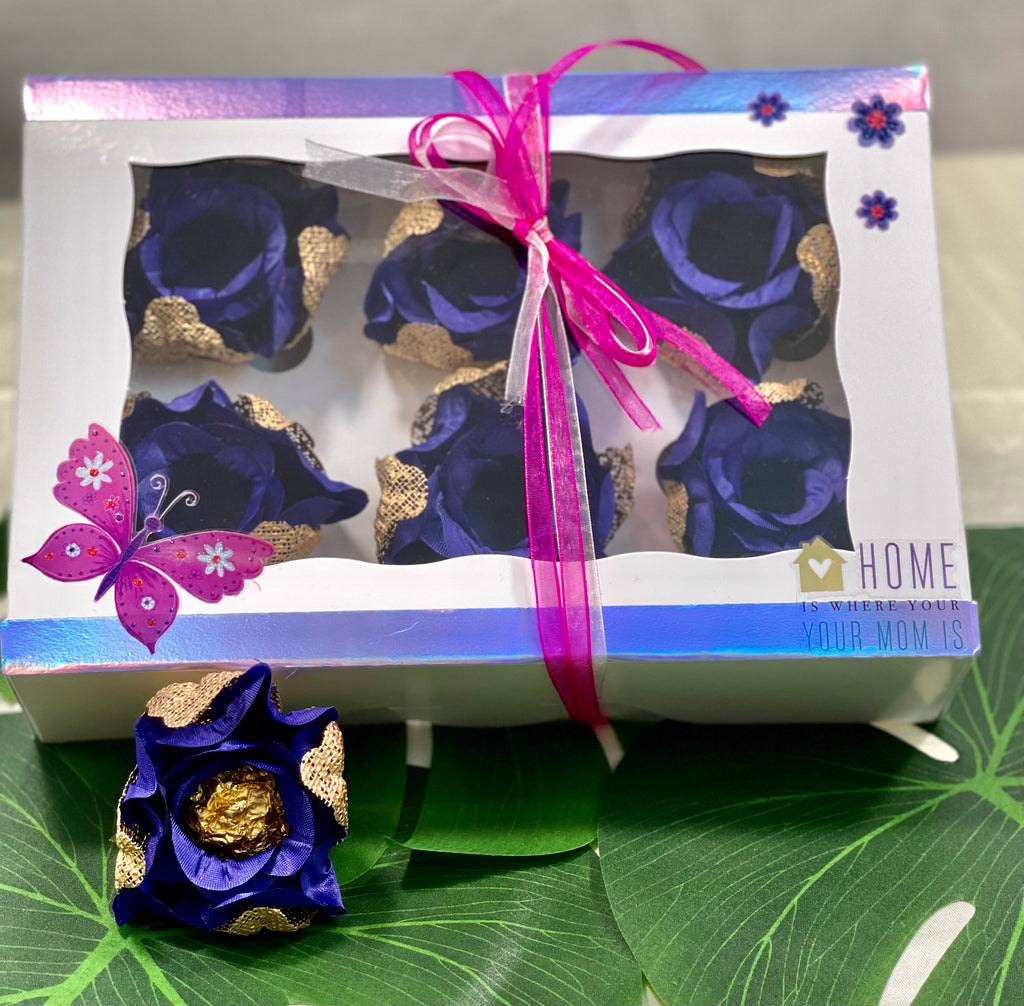 The image shows a box of 6 royal blue and gold rosebuds. The box is decorated with a pink butterfly and flowers. there is also a sticker saying "Home is where your mom is". There is one rose holding a gold chocolate truffle place in front of the box.