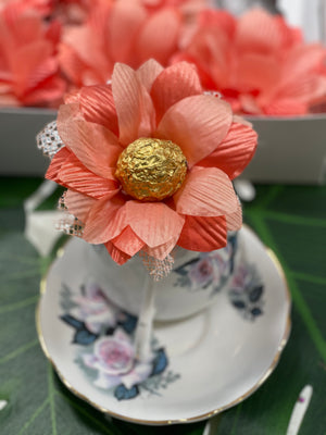 The image shows a coral color flowers sitting on a teacup and saucer. The flower holds a chocolate wrapped in gold. This flower is a truffle holder