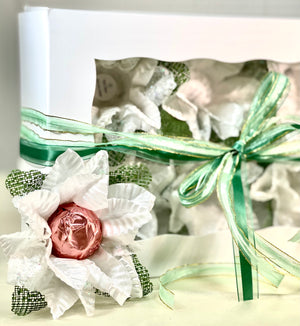 Image of a white box with green ribbon. The box comes with 6 flowers. These flowers are white with green accents. Each flower has a little cup in the middle which is used to hold sweets. Theses flowers are sweet holder. There is 1 flower sitting in front of the box. In the flower there is a chocolate truffle wrapped in rose gold foil.